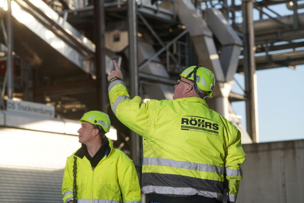 Zwei Männer in gelben Schutzjacken und Helmen stehen bei einer Industrieanlage, einer zeigt nach oben